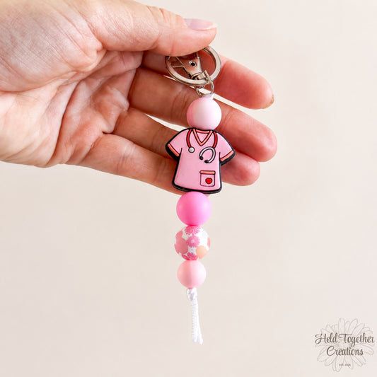 Hand holding a keychain with a pink stethoscope and scrubs design on a beige background.
