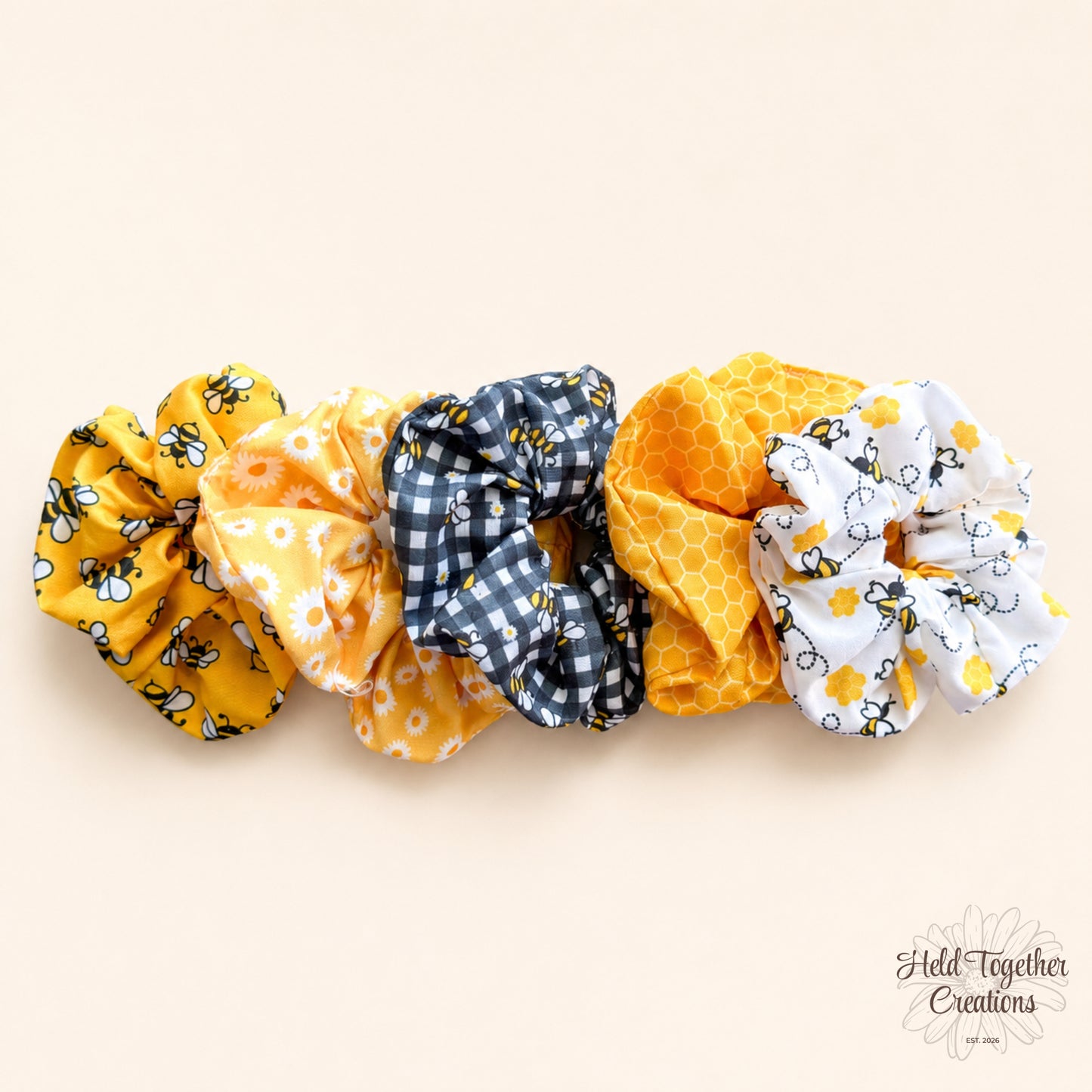 Five handmade Honey Bloom scrunchies in yellow, white, black and grey bee, daisy, gingham and honeycomb prints on a warm ivory background.