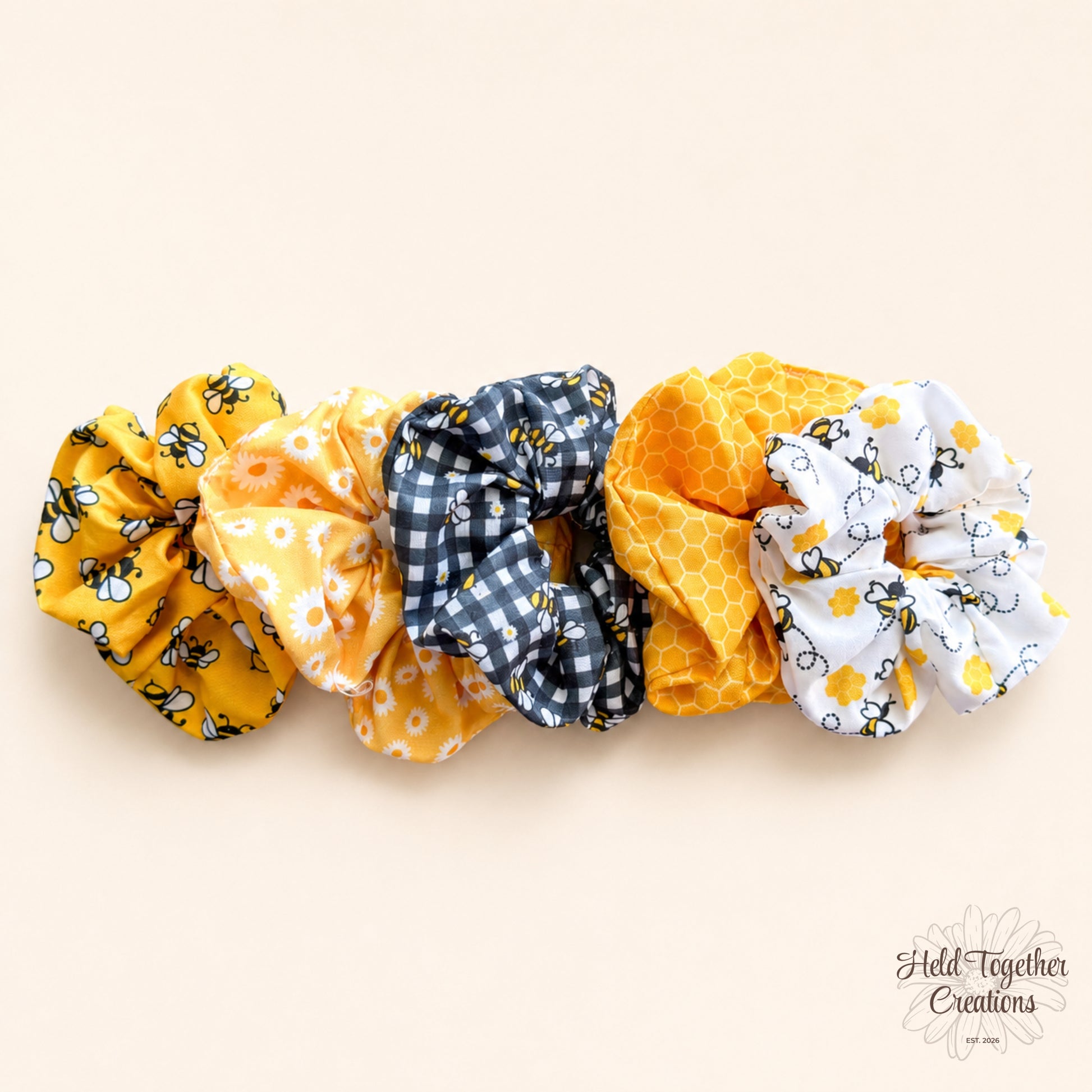 Five handmade Honey Bloom scrunchies in yellow, white, black and grey bee, daisy, gingham and honeycomb prints on a warm ivory background.