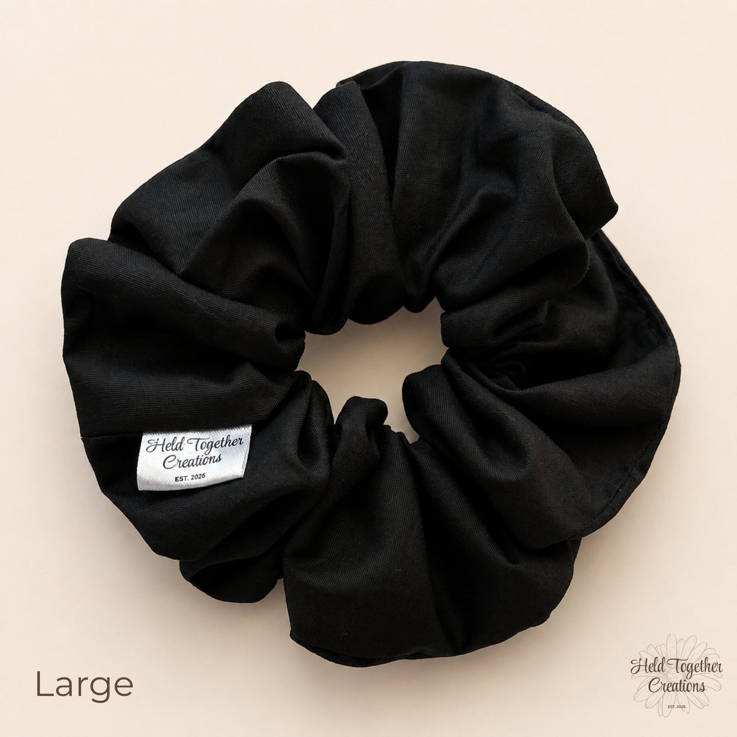 A large black fabric scrunchie with a white brand label reading 'Held Together Creations' on a light beige background. The word 'Large' is printed in the lower-left corner, and the brand logo appears in the lower-right corner.