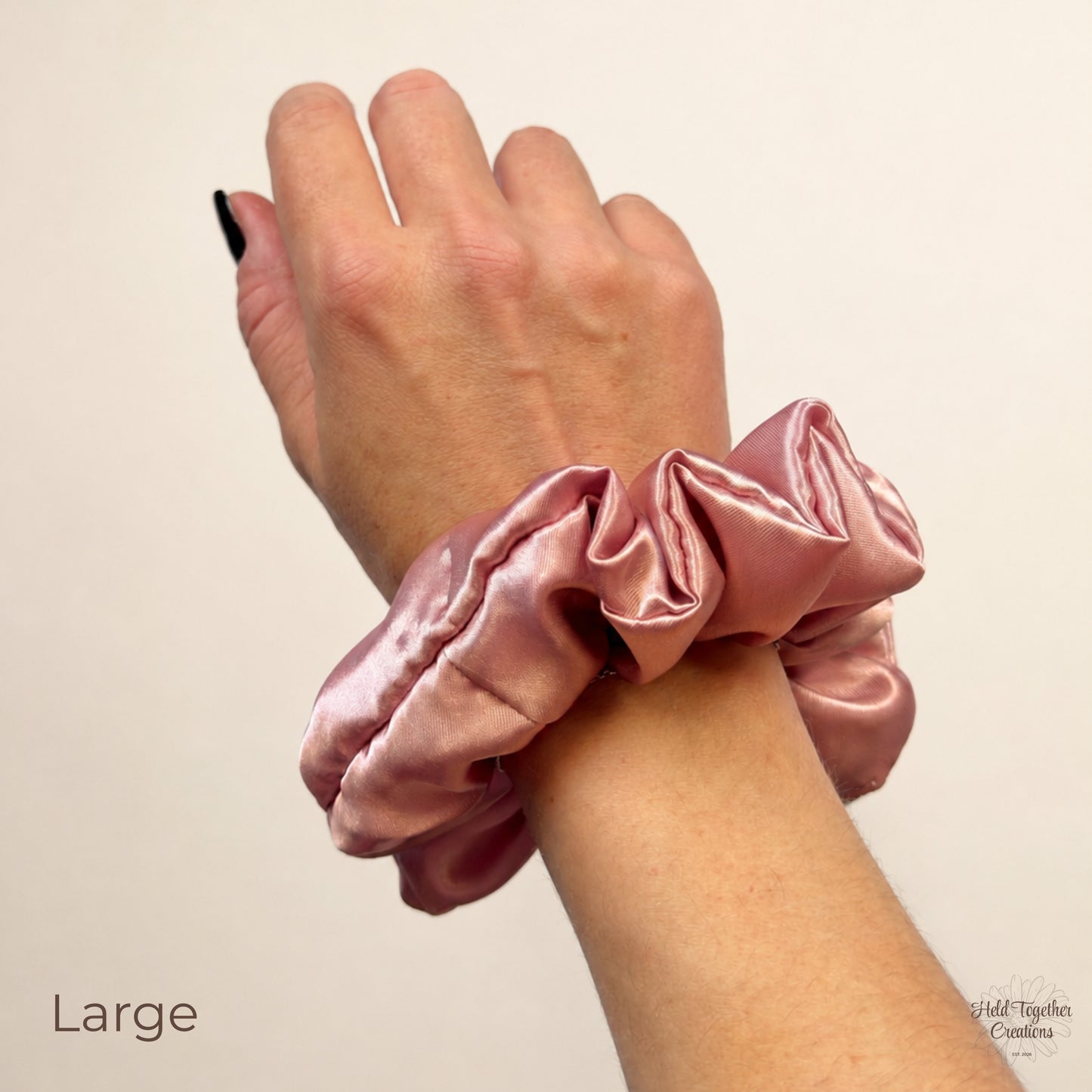 A dusty pink satin scrunchie worn on a person's wrist, shown against a plain light beige background with the word 'Large' in the lower left corner.