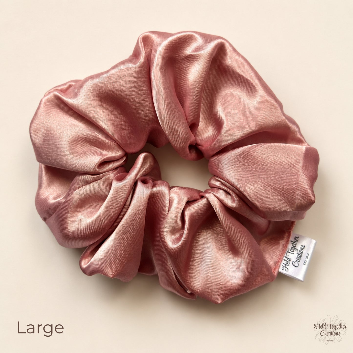 A large satin hair scrunchie in a dusty pink colour, displayed on a light beige background with a small white brand tag on the side.