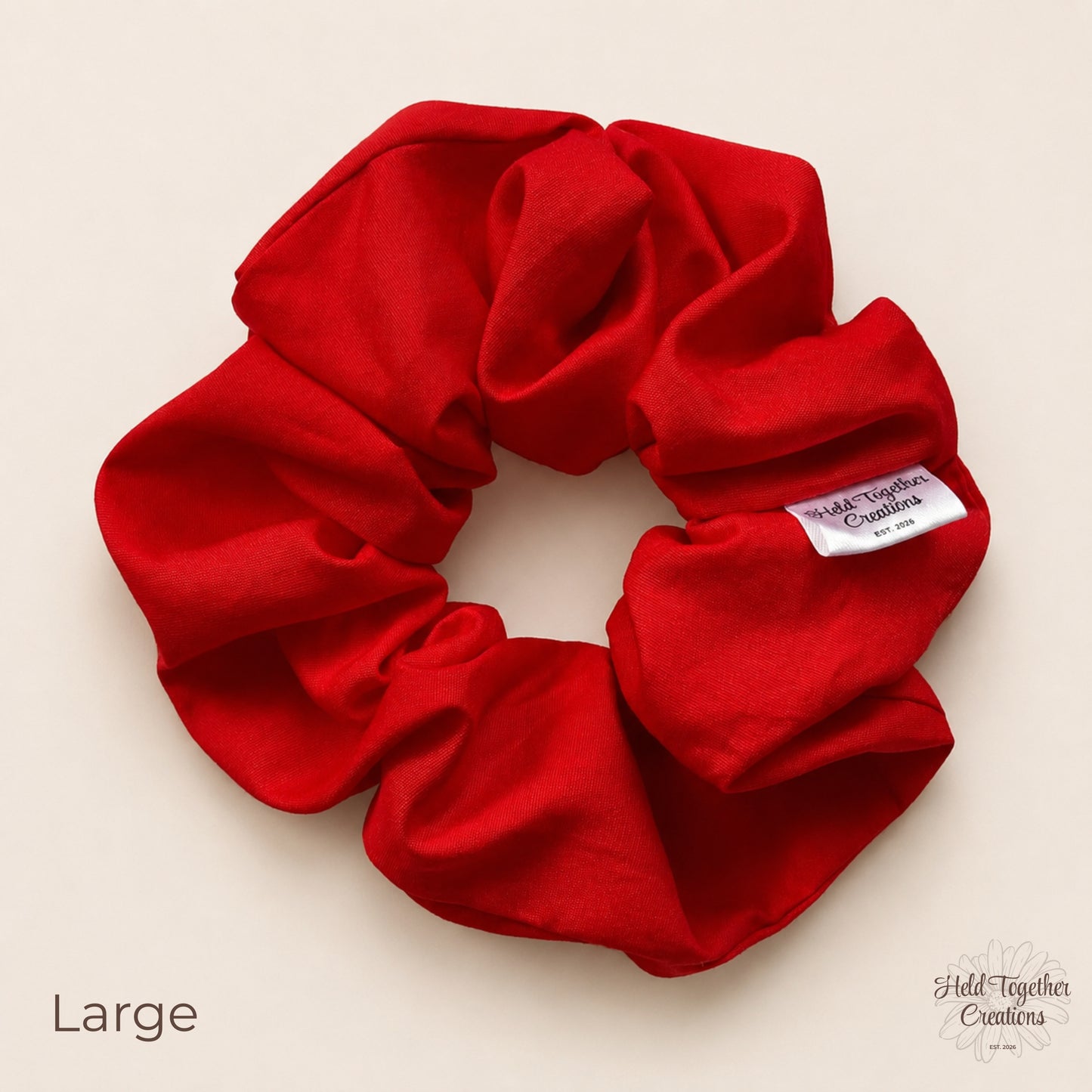 Large red cotton scrunchie laid flat on a warm ivory background with branded tag visible.