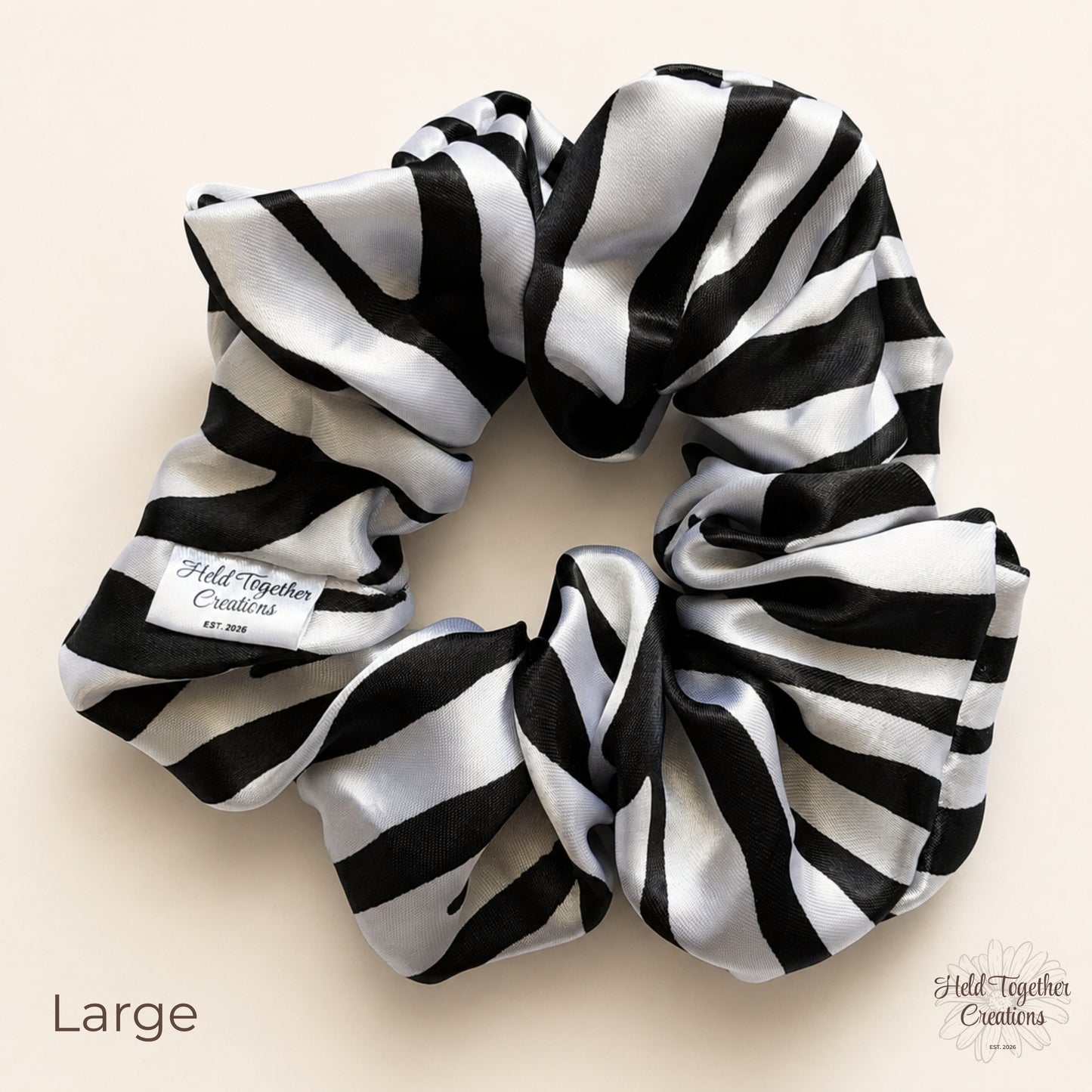 A large black and white striped satin scrunchie with a white paper label reading 'Held Together Creations Est. 2026' on a light beige background.