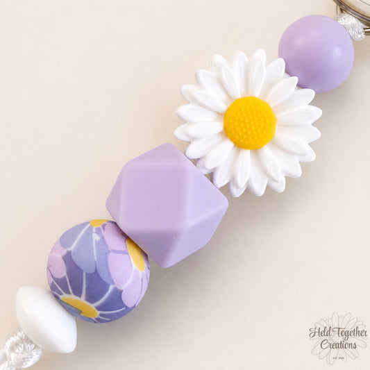 Lilac daisy silicone bead keyring with white daisy bead, lilac beads and silver keyring hardware on a soft neutral background.