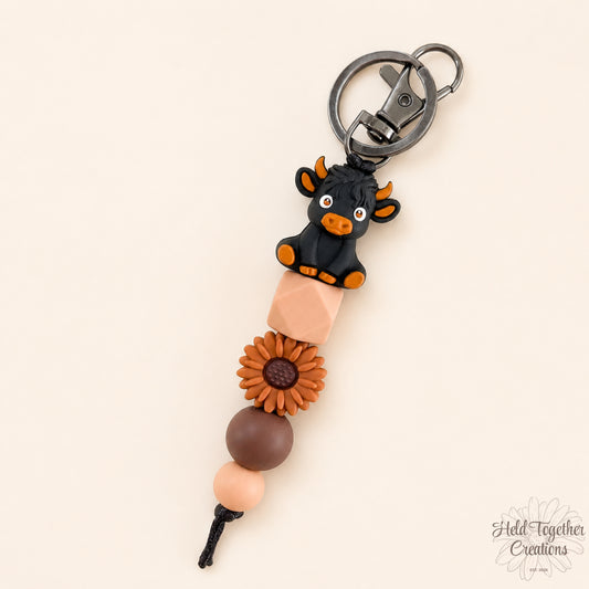 Keychain with black cow figure, brown flower, and beige beads on a white background