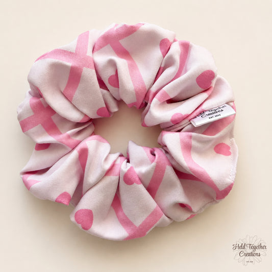 A fabric hair scrunchie with a pink and white heart and stripe pattern, shown on a light beige background. A small white brand tag is visible on the right side of the scrunchie.