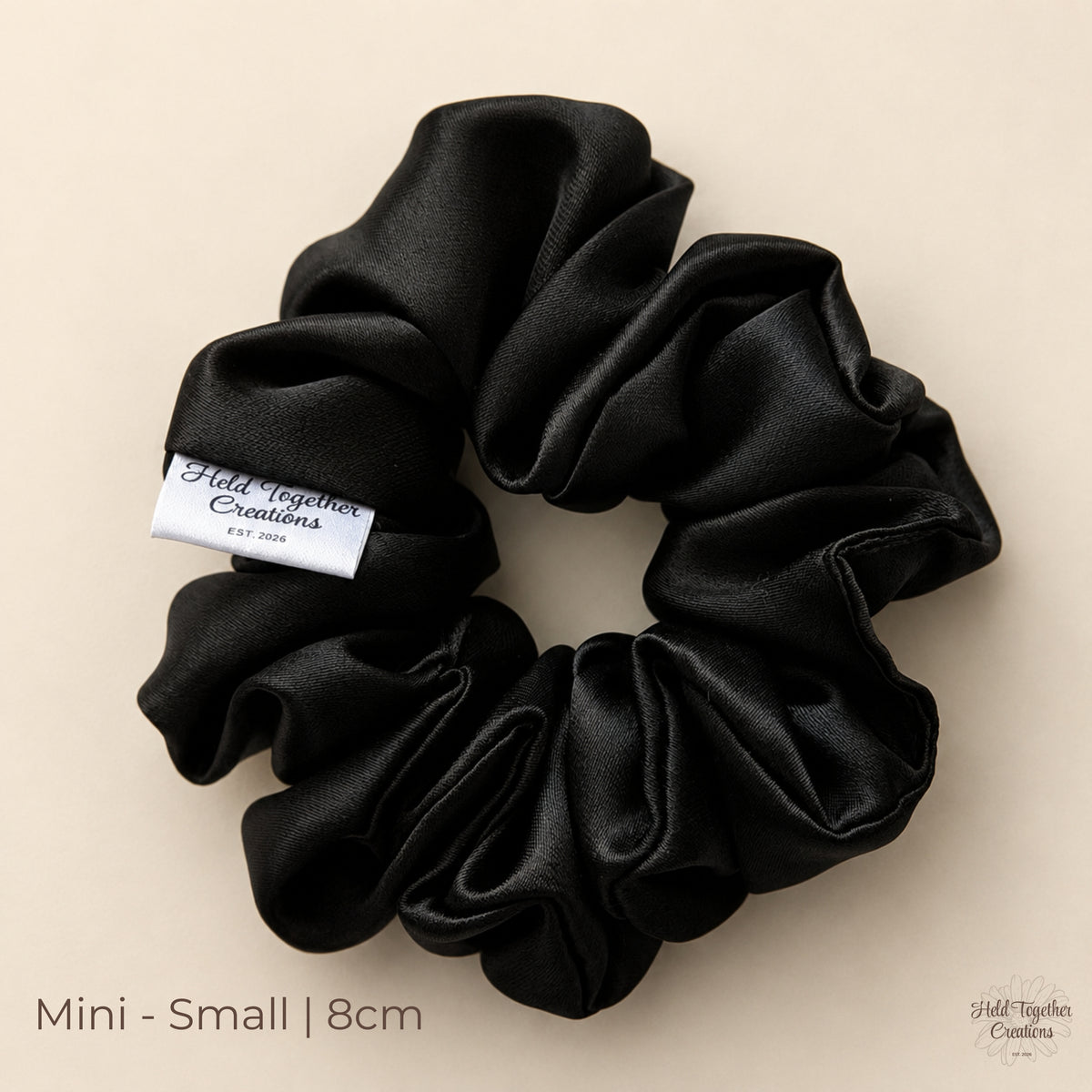 Black satin Mini scrunchie (8cm) laid flat on a warm ivory background, showing soft gathered folds and Held Together Creations label.