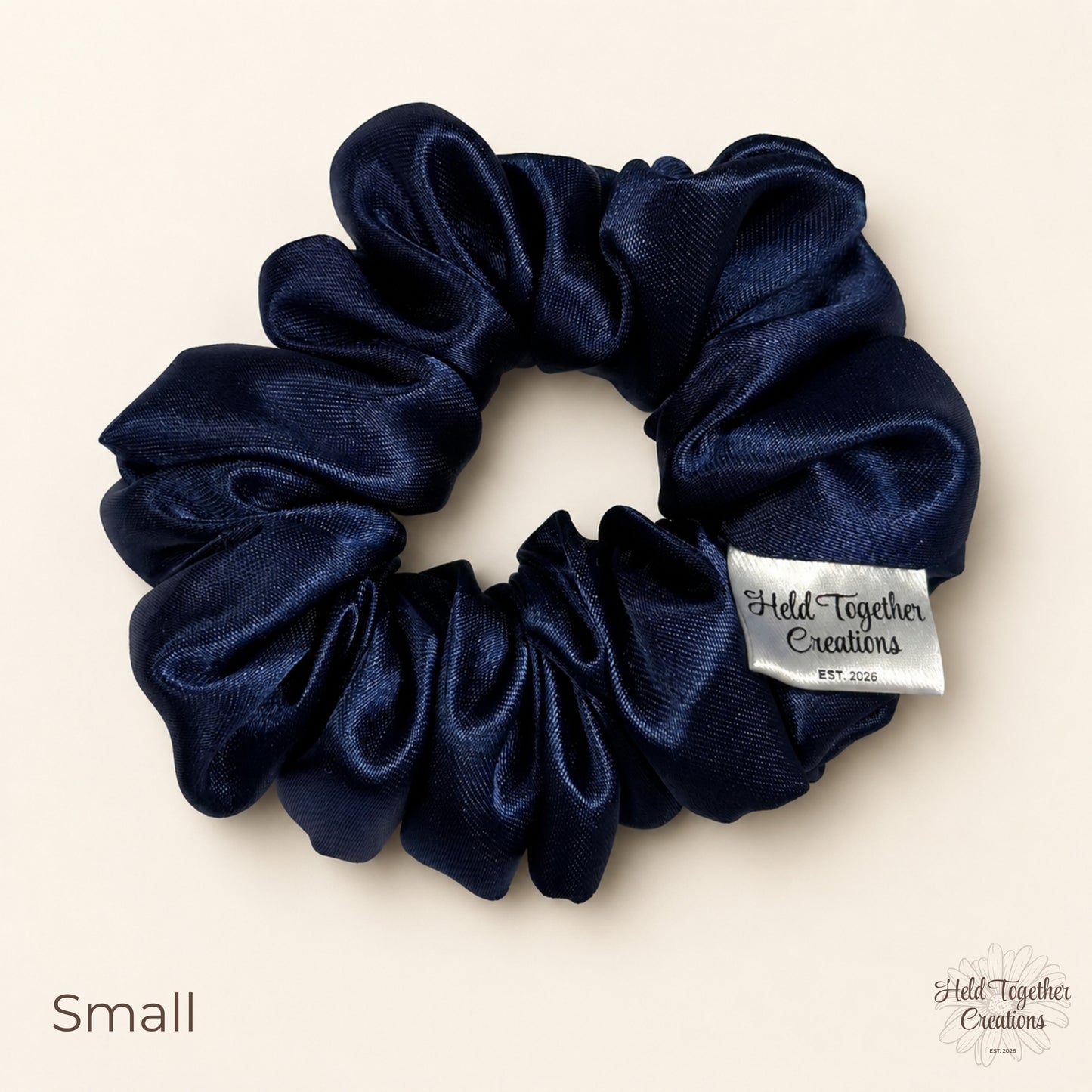 Small navy satin scrunchie photographed flat on a neutral studio background with brand label visible.