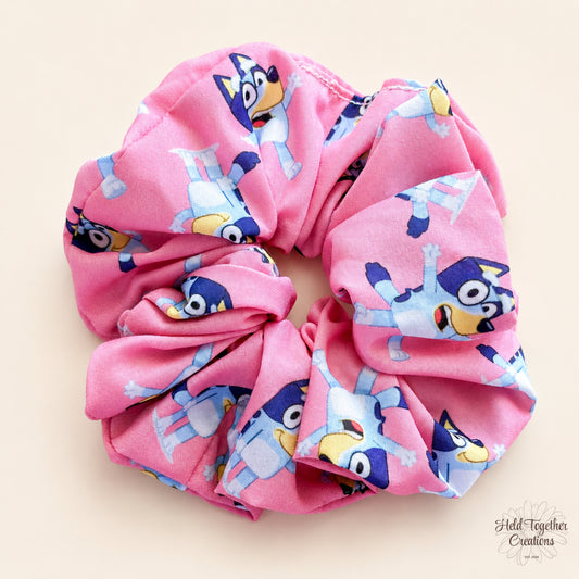 Bright pink Party Pup Scrunchie laid flat on a warm ivory background, showing its colourful cartoon print.