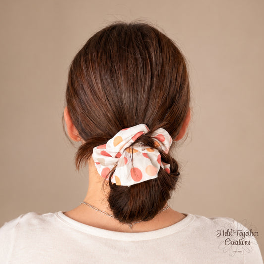 White handmade scrunchie with peach, coral, pink and neutral spot pattern by Held Together Creations.