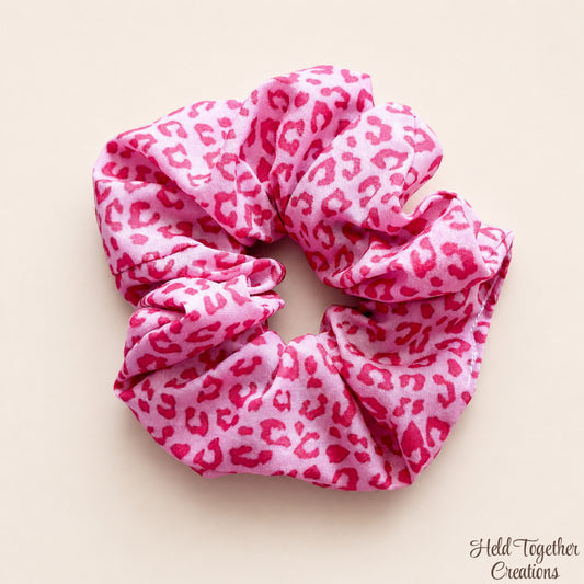 A pink fabric scrunchie with a darker pink leopard print pattern, photographed on a solid light beige background. A watermark in the bottom right corner reads 'Held Together Creations'.