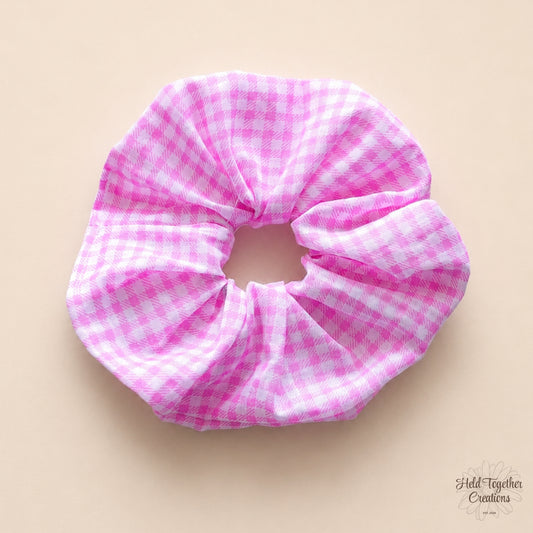 Handmade pink and white gingham fabric scrunchie displayed on a warm neutral background.