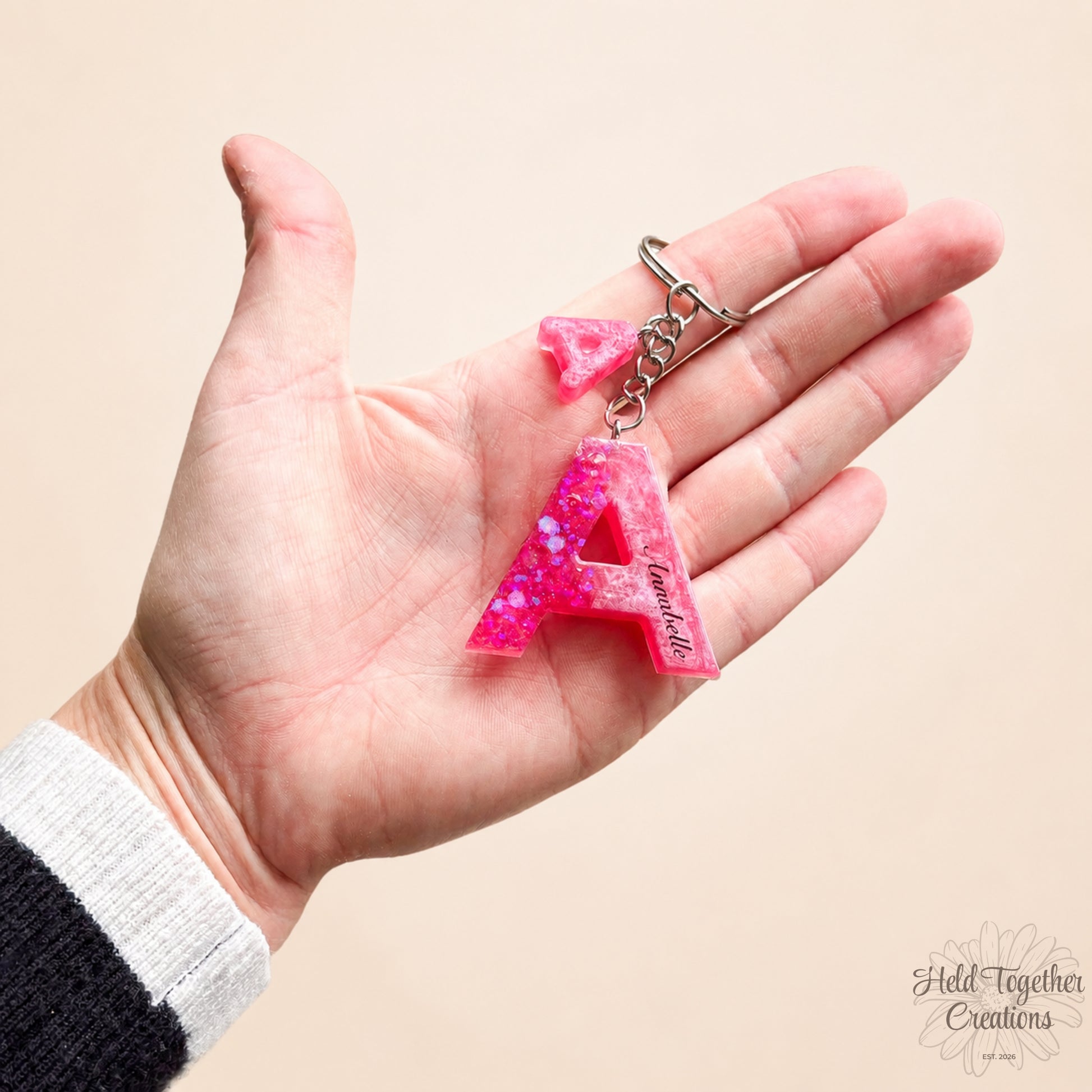 Hand holding a pink glittery letter 'A' keychain with 'Annabelle' engraved on a beige background.