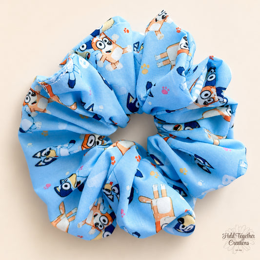 A light blue fabric scrunchie featuring a cartoon dog print, displayed on a solid beige background.