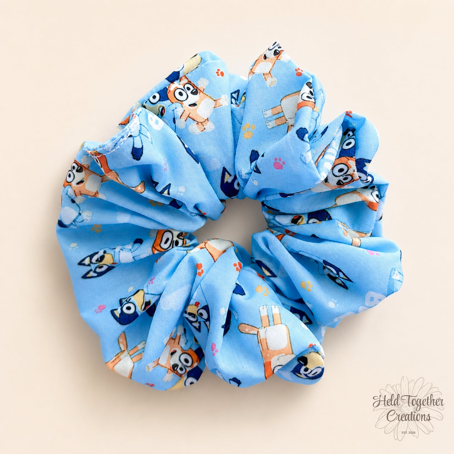 Playdate Blue handmade scrunchie in bright blue puppy-print fabric on a warm ivory background with Held Together Creations branding.