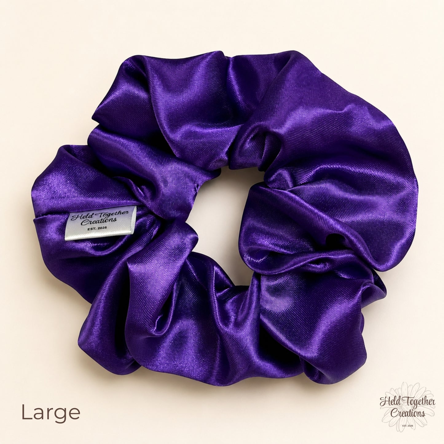 Large purple satin scrunchie laid flat on a warm ivory background, showing the shiny satin fabric and Held Together Creations label.