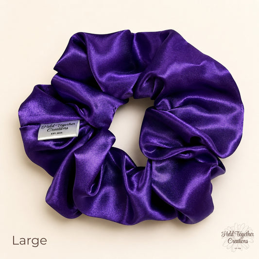 Large purple satin scrunchie laid flat on a warm ivory background, showing the shiny satin fabric and Held Together Creations label.