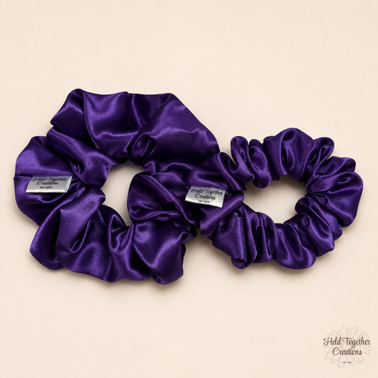 Small and large purple satin scrunchies displayed together on a warm ivory background, showing the size difference and glossy satin finish.