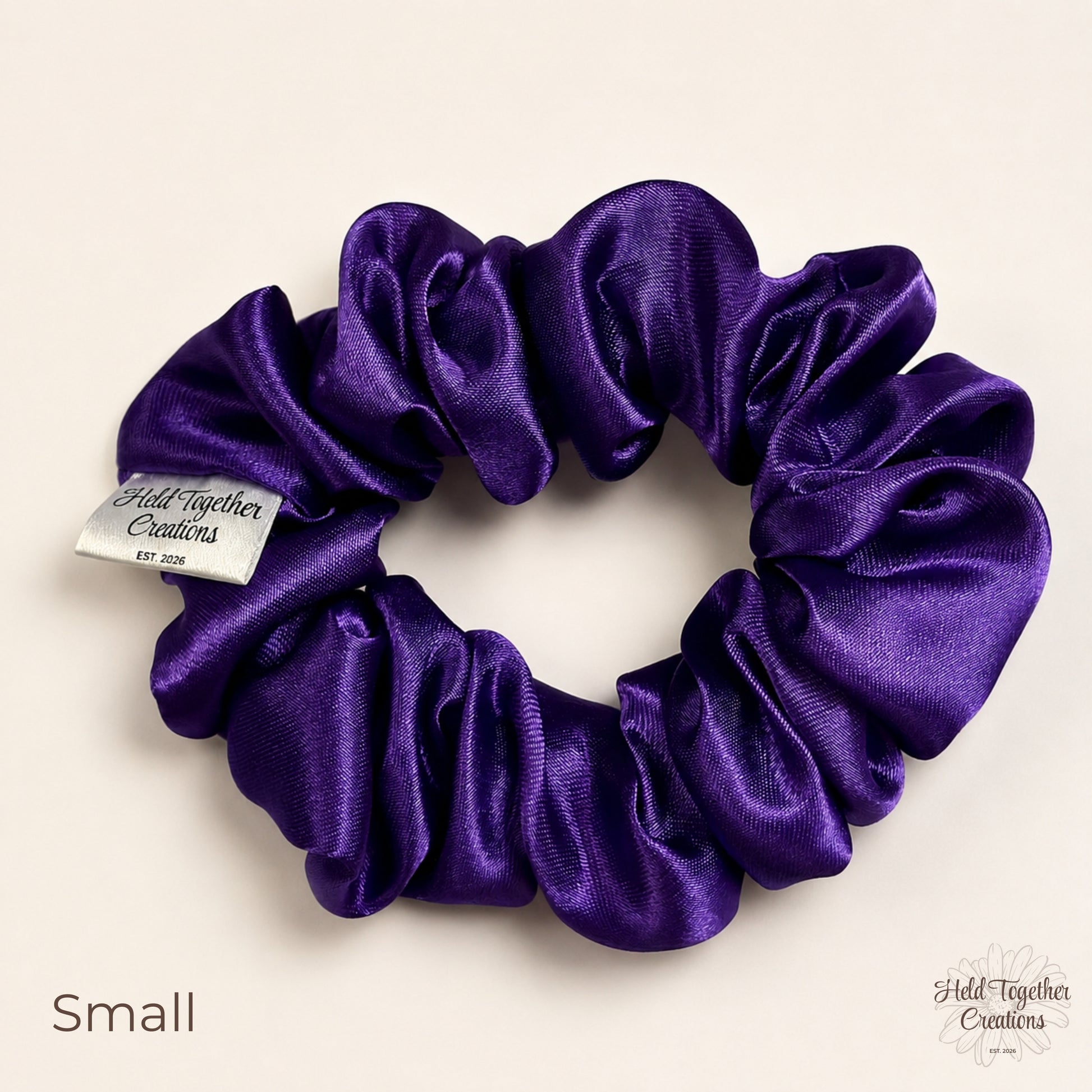 Small purple satin scrunchie laid flat on a warm ivory background with the Held Together Creations label visible.