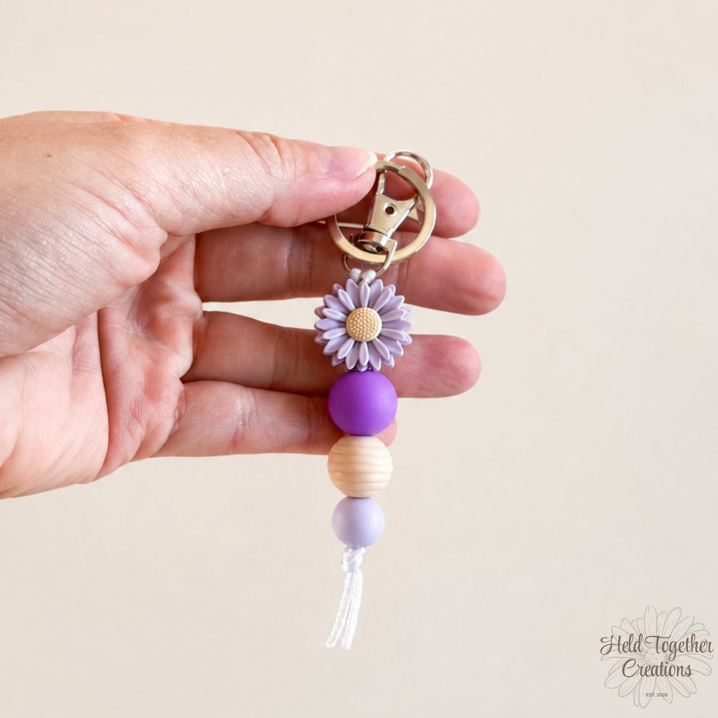 Hand holding a keychain with purple beads and a flower charm against a beige background