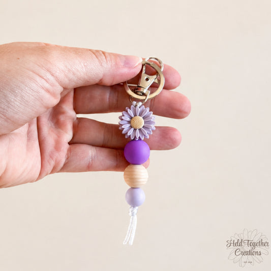 Hand holding a keychain with purple beads and a flower charm against a beige background