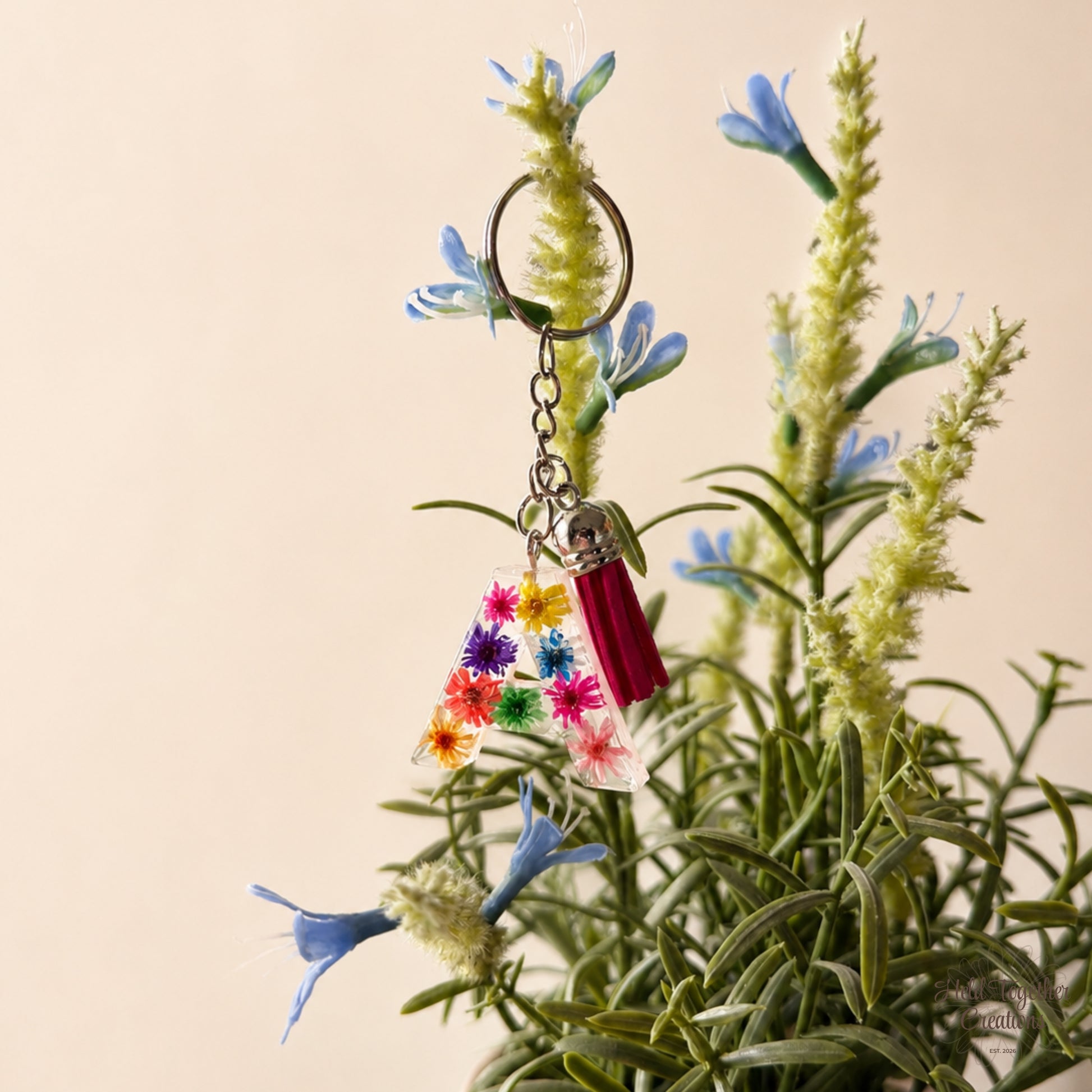 A decorative keychain with a silver key ring, chain, small silver bell, pink tassel, and a clear resin letter A filled with colourful flower shapes, hanging in front of artificial blue flowers and green stems against a warm beige background.