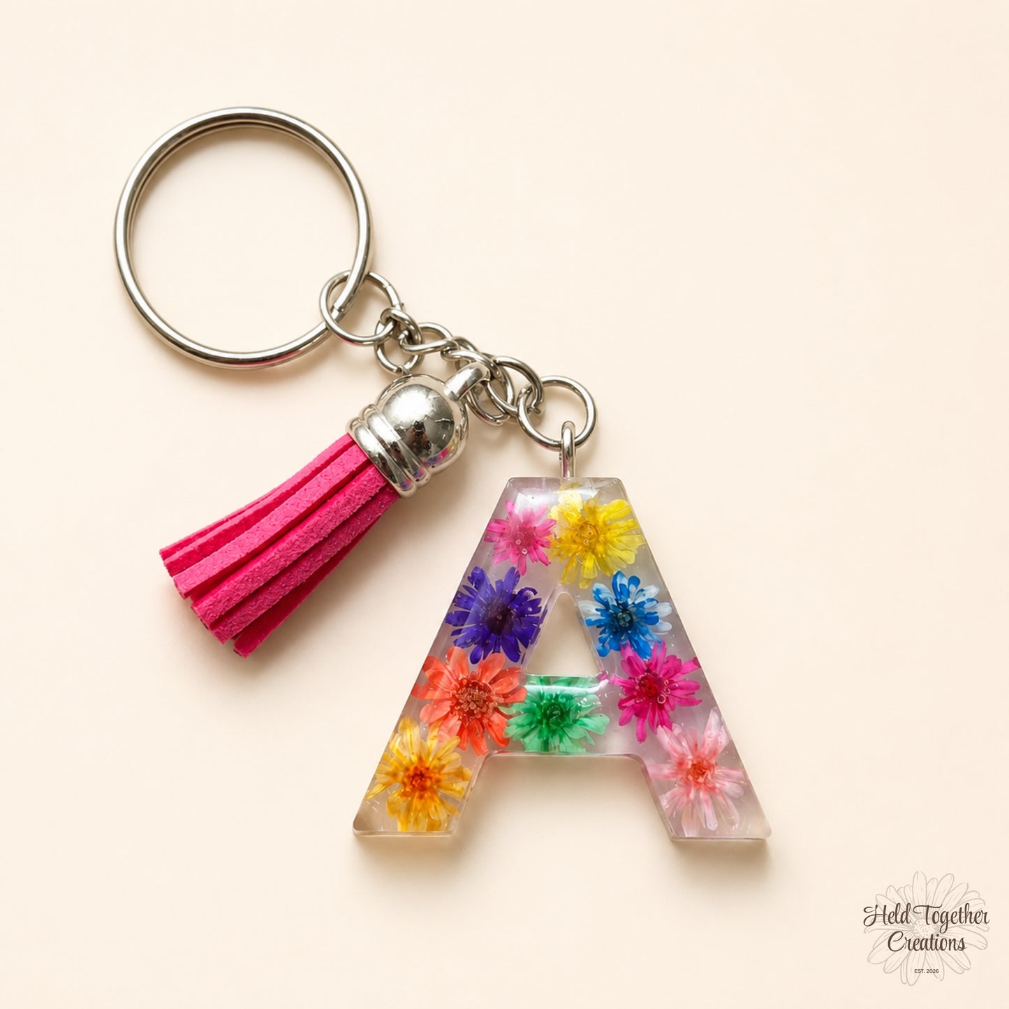 A silver metal keychain featuring a clear resin letter 'A' filled with colourful dried flowers, a bright pink tassel, and a silver key ring, set against a solid pale peach background.