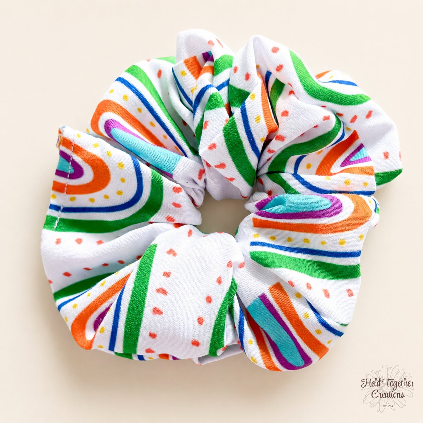 Rainbow Rush Scrunchie laid flat on a warm ivory background with bright rainbow stripes and dot details.