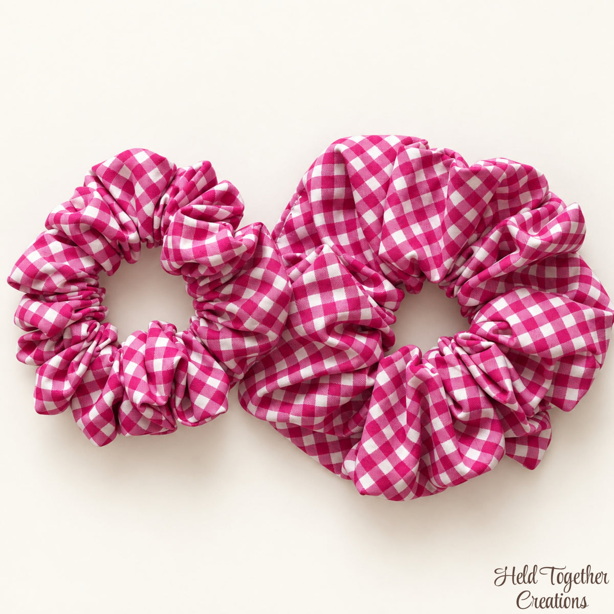 Two pink and white gingham fabric scrunchies on a light cream background, with a 'Held Together Creations' watermark in the bottom right corner.