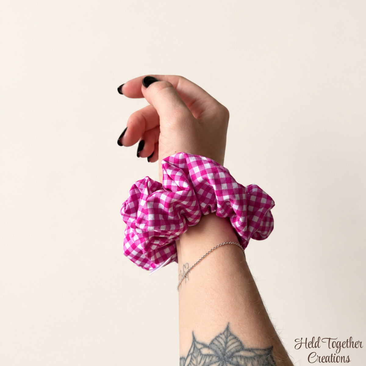 A pink and white gingham fabric scrunchie worn on a person's wrist, shown against a plain light beige background. The person's hand and forearm are visible, along with a silver chain bracelet and a tattoo on the forearm.