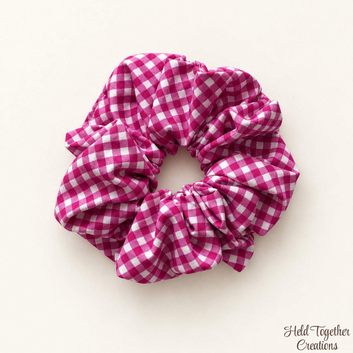 A fabric hair scrunchie with a pink and white gingham check pattern, photographed on a light cream background with a small 'Held Together Creations' watermark in the bottom right corner.