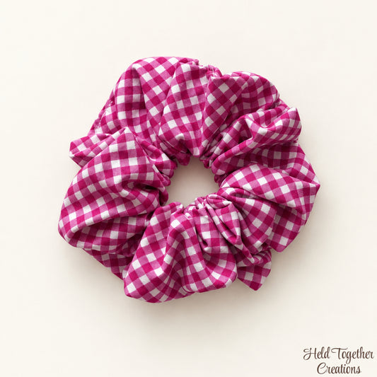A fabric hair scrunchie with a pink and white gingham check pattern, photographed on a light cream background with a small 'Held Together Creations' watermark in the bottom right corner.