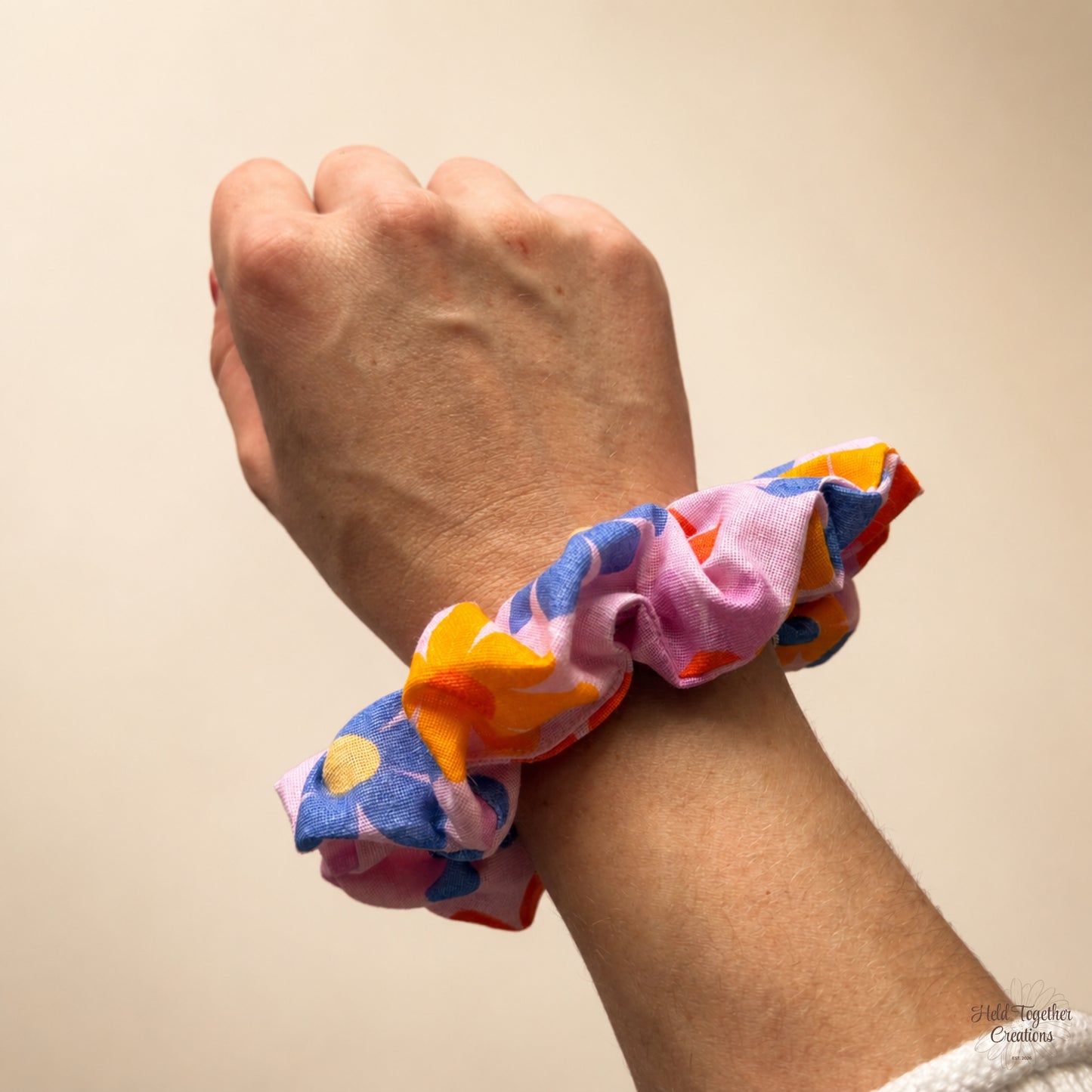 A close-up of a person's wrist wearing a fabric scrunchie with a colourful floral pattern in pink, blue, orange, and yellow, set against a plain beige background.