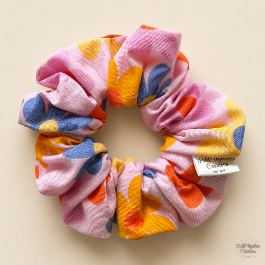 A fabric scrunchie with a retro floral pattern in pink, orange, yellow, and blue, featuring a white brand tag that reads 'Held Together Creations Est. 2026' on a light beige background.