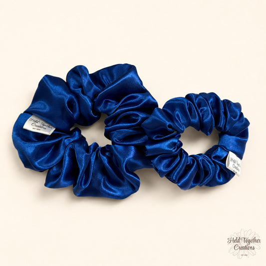 Small and large royal blue satin scrunchies laid side by side on a warm ivory background.