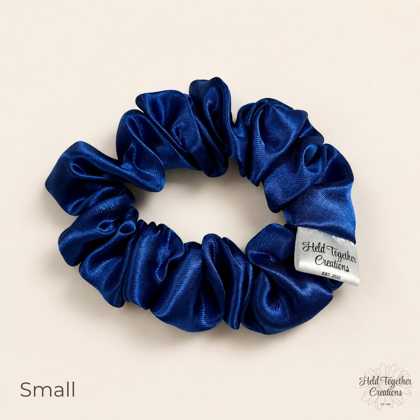 Small royal blue satin scrunchie laid flat with glossy gathered satin fabric and brand tag.