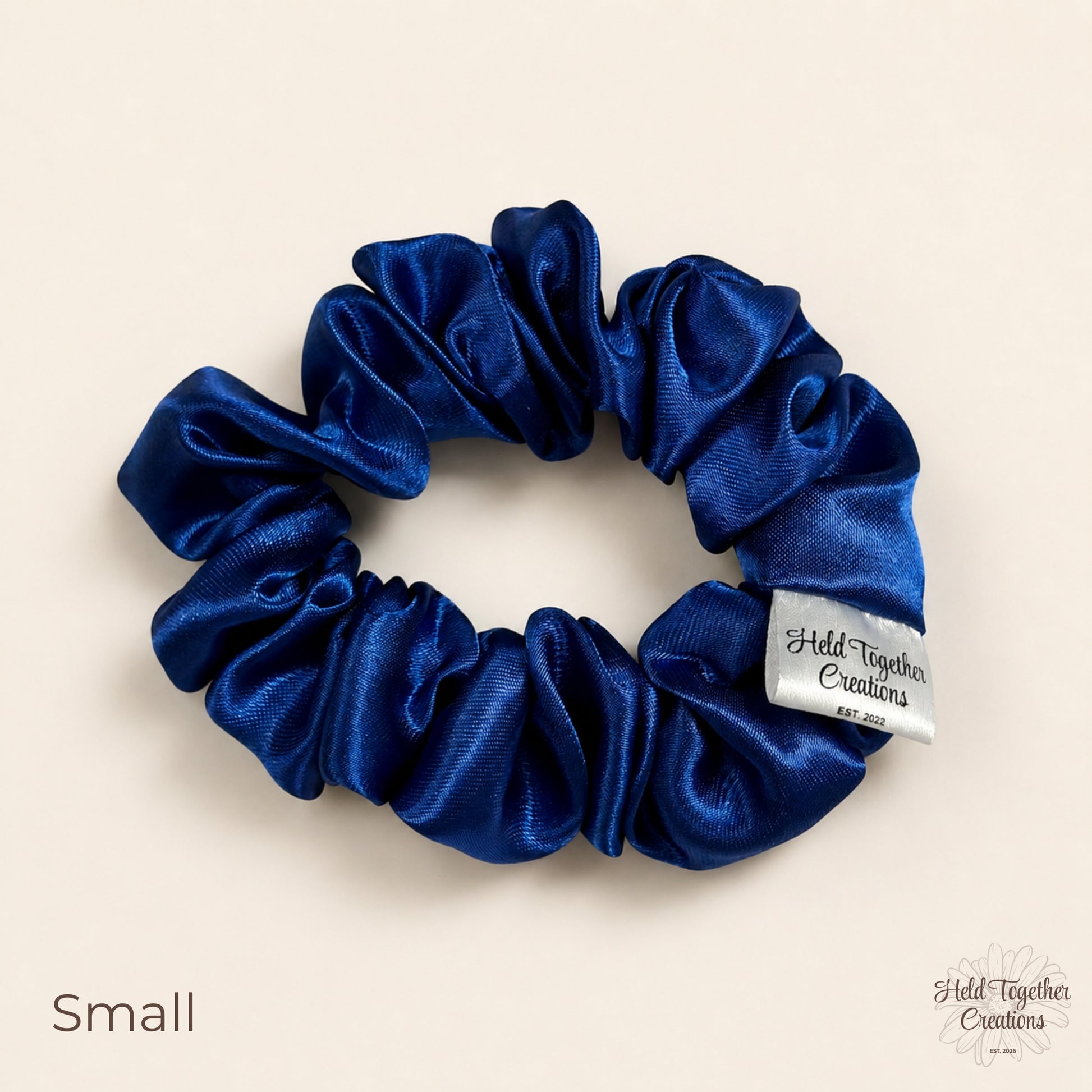 Small royal blue satin scrunchie laid flat with glossy gathered satin fabric and brand tag.