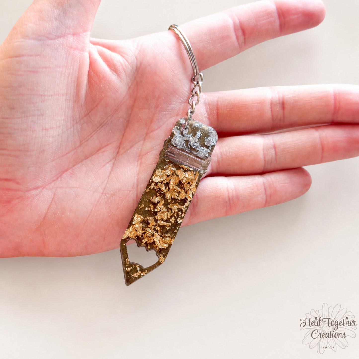 Handmade pencil-shaped resin keyring with gold and silver flakes and metal keyring hardware on a soft neutral background.