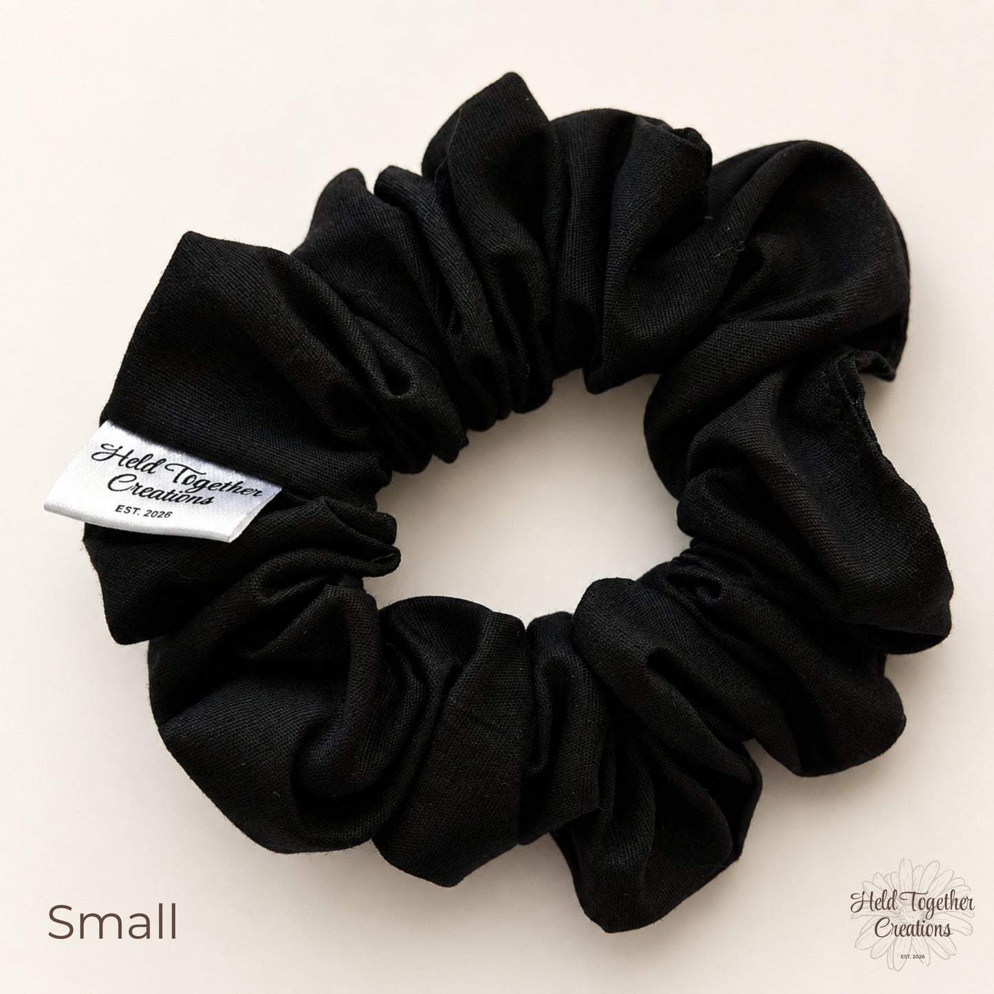 A black cotton scrunchie with a white sewn-in label that reads 'Held Together Creations EST. 2026' on a light beige background.