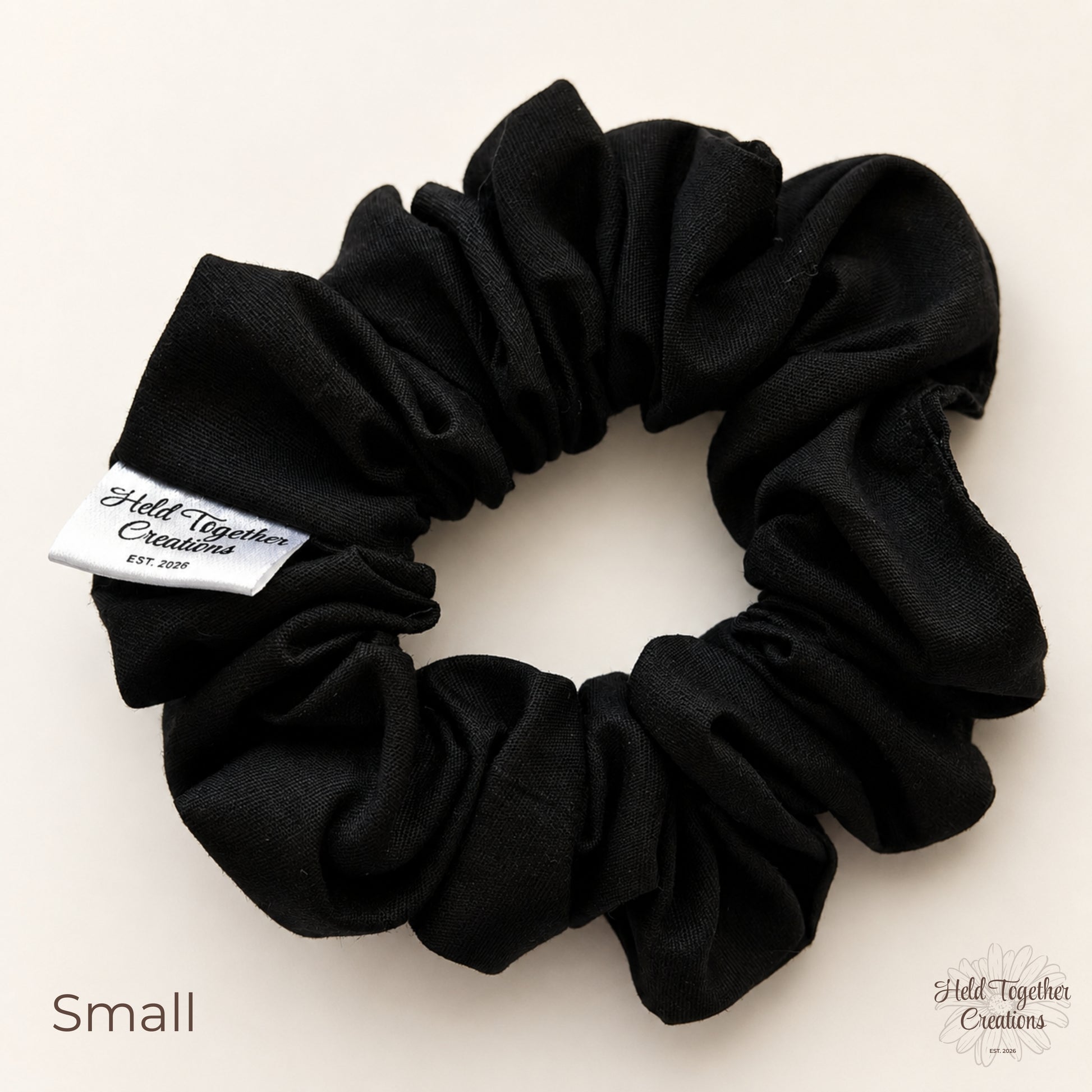 A black cotton scrunchie with a white sewn-in label that reads 'Held Together Creations EST. 2026' on a light beige background.