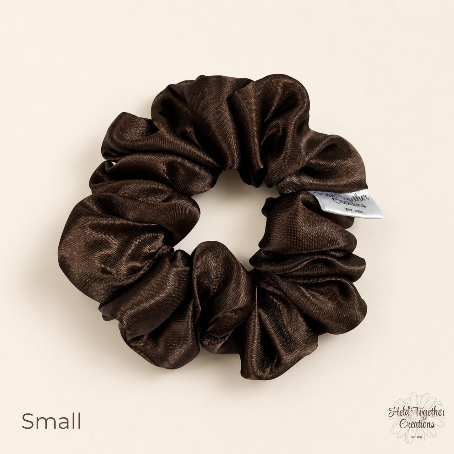 Small brown satin scrunchie on a warm ivory background with soft gathered satin fabric and HTC label.