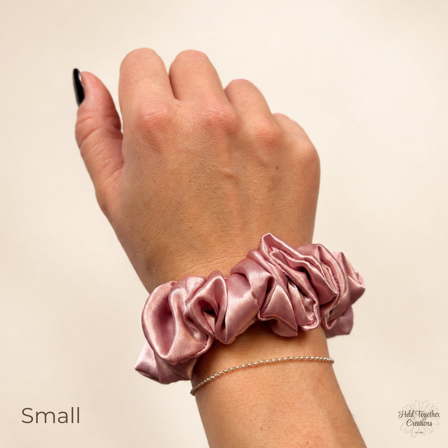 A close-up of a person's hand and wrist wearing a dusty pink satin scrunchie bracelet. A thin silver chain bracelet is also visible underneath.