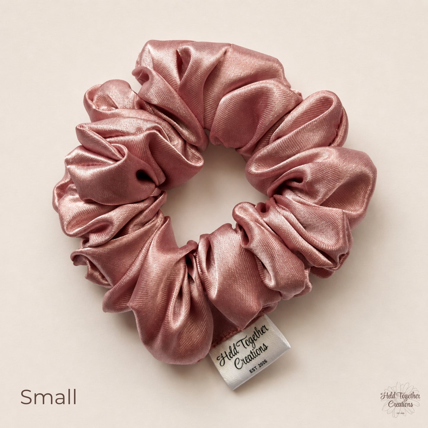 A dusty pink satin scrunchie with a white brand tag reading 'Held Together Creations' on a warm beige background. The word 'Small' is printed in the bottom-left corner.