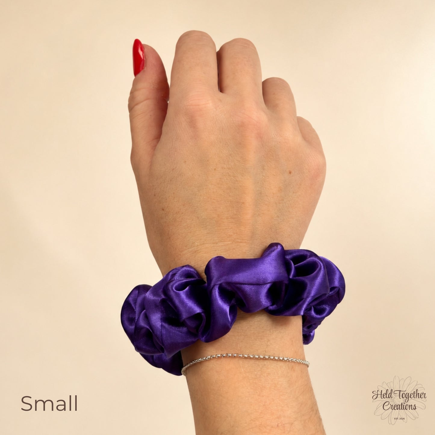Small purple satin scrunchie worn on a wrist, showing the compact gathered satin style against a warm ivory background.