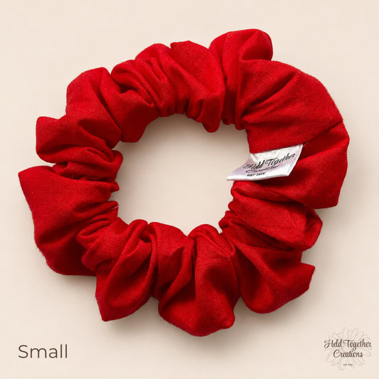 Small red cotton scrunchie laid flat on a warm ivory background with branded tag visible.