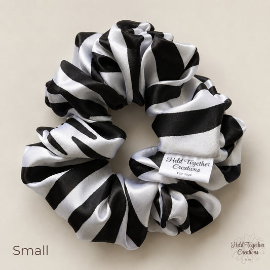 A black and white striped satin hair scrunchie with a white fabric tag reading 'Held Together Creations Est. 2026' on a light beige background.
