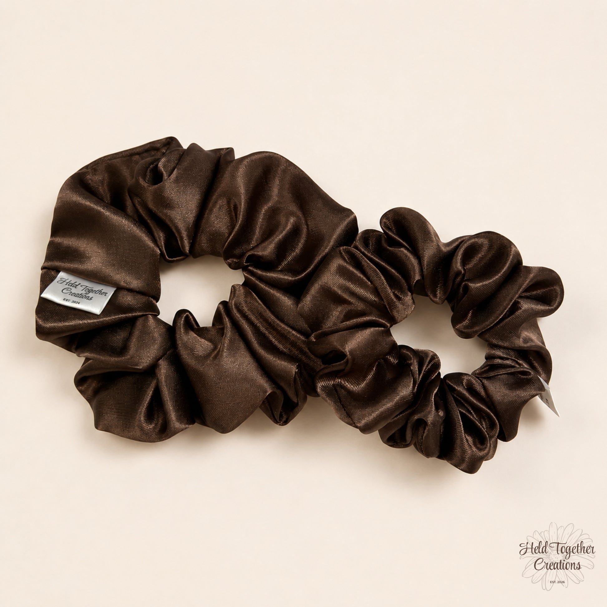 Small and large brown satin scrunchies displayed together on a warm ivory background showing size difference.