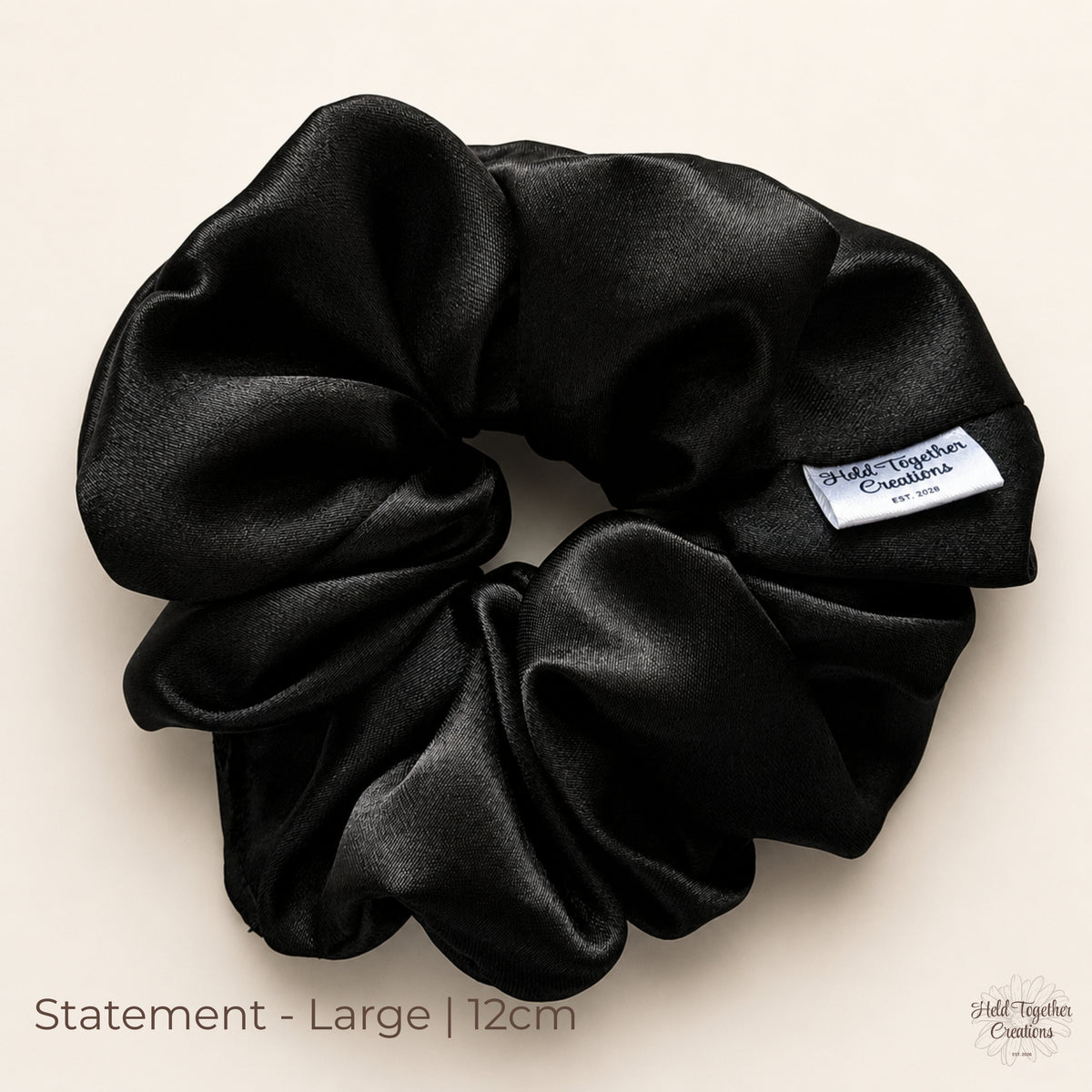 Black satin Statement scrunchie (12cm) laid flat on a warm ivory background, showing full ruched volume and Held Together Creations label.