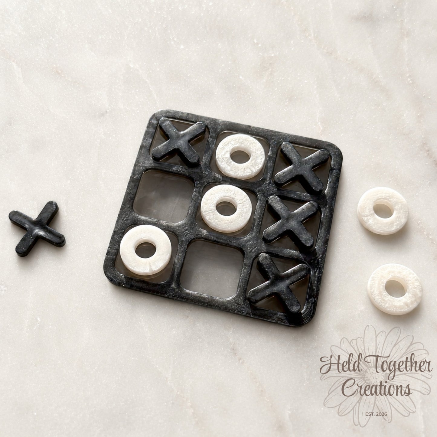 Handmade monochrome resin tic-tac-toe game with black board, black X pieces and white O pieces on a soft neutral background.